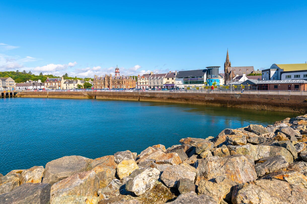 View of Stornoway’s harbor in Scotland featuring calm blue water, rocky shoreline and a row of historic buildings including a clock-topped stone hall and a tall church spire.