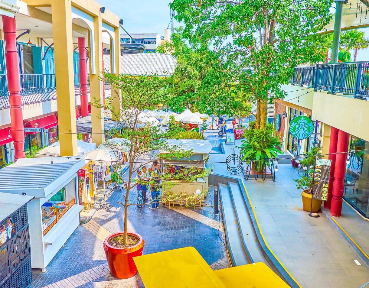 Outdoor shopping area in Bangkok featuring small market stalls, umbrella-shaded vendors and vibrant storefronts with trees and greenery.