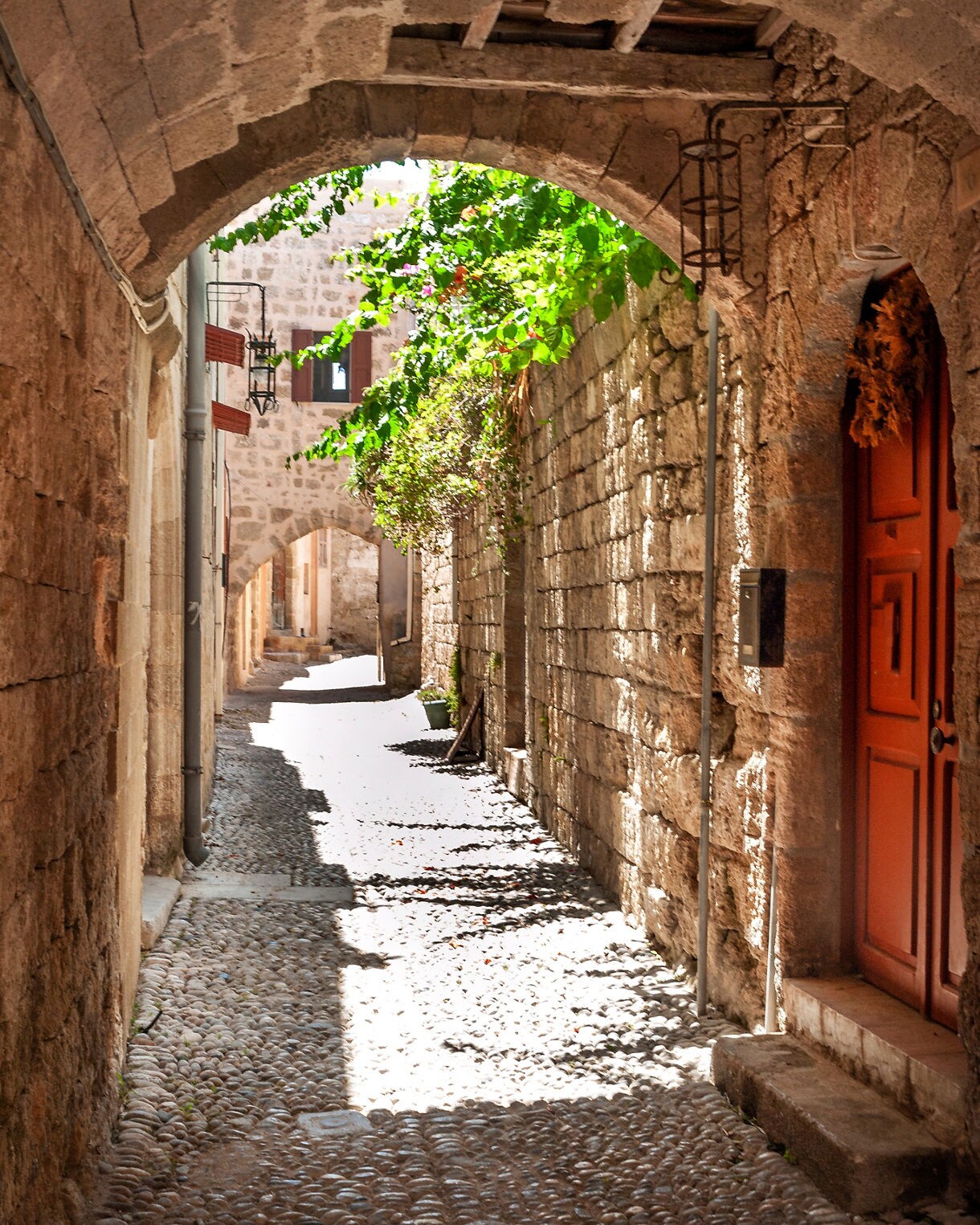 Sunlit cobblestone alley in Rhodes Old Town framed by stone arches, climbing vines and wooden doors.