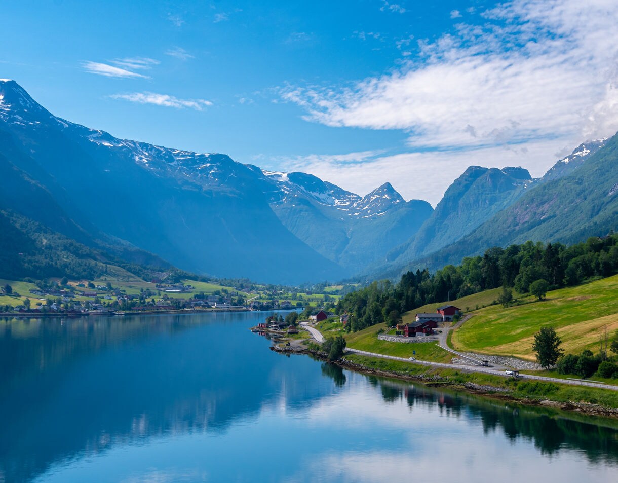 Stryn Lake in Norway with calm reflective water, green fields, red farmhouses and surrounding snow-capped mountains under a blue sky.