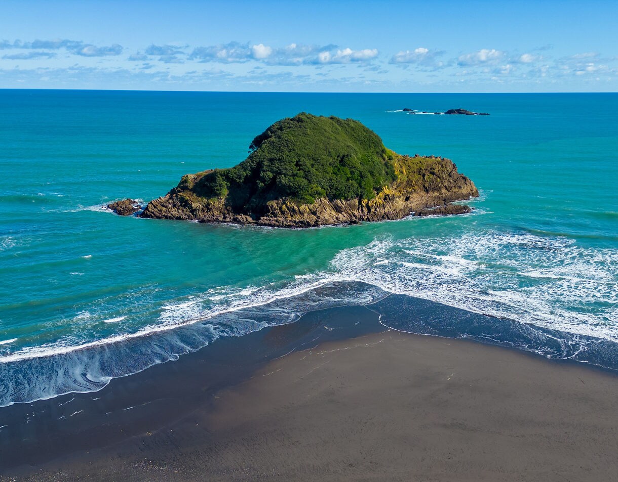 A small rocky island covered in dense greenery sits just offshore, surrounded by turquoise waves and a black sand beach in New Plymouth.