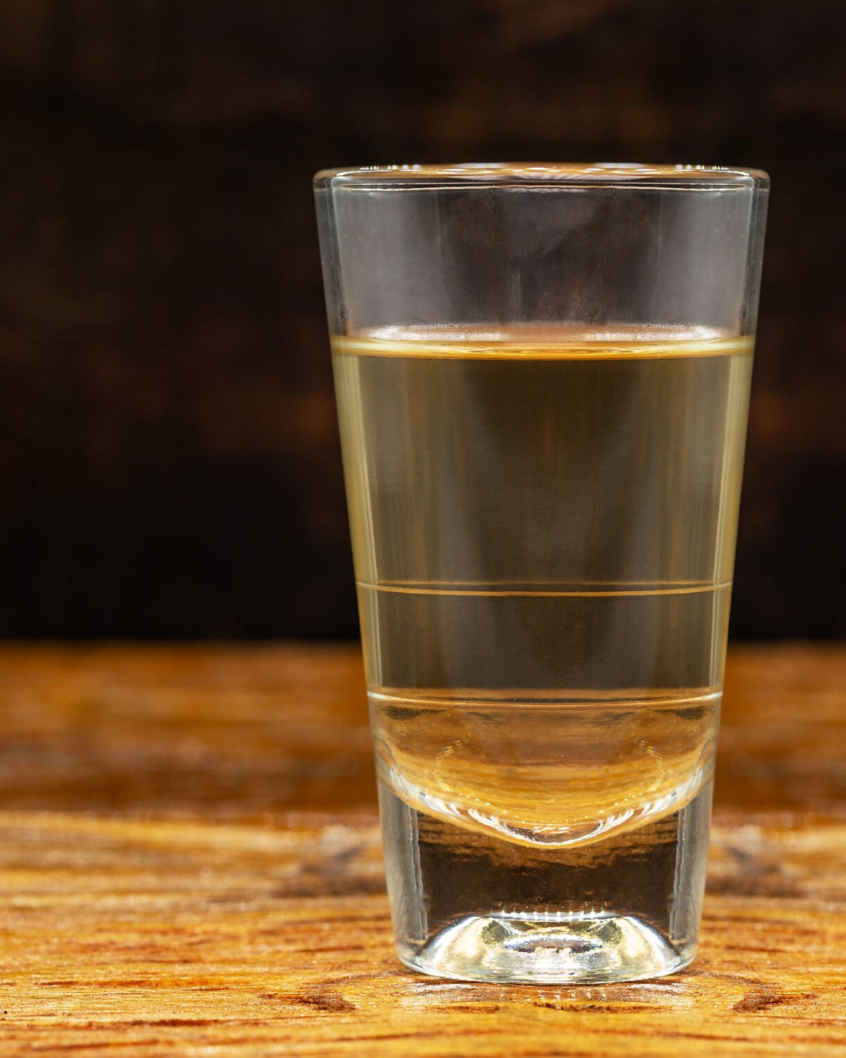 Close-up of a clear shot glass filled with pale sugarcane liquor on a rustic wooden surface.