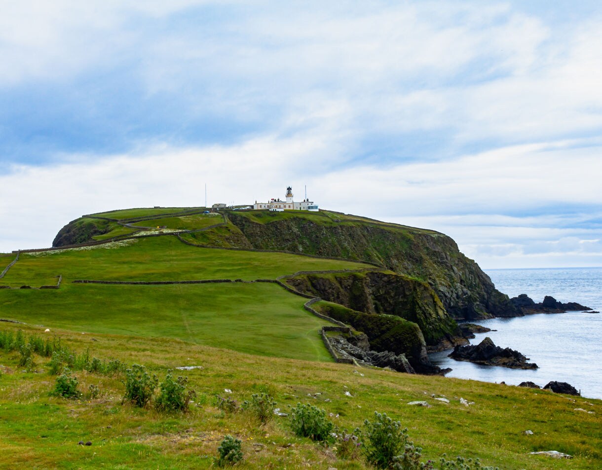 Distant view of Sumburgh Head lighthouse perched on grassy cliffs overlooking the rocky shoreline and sea.