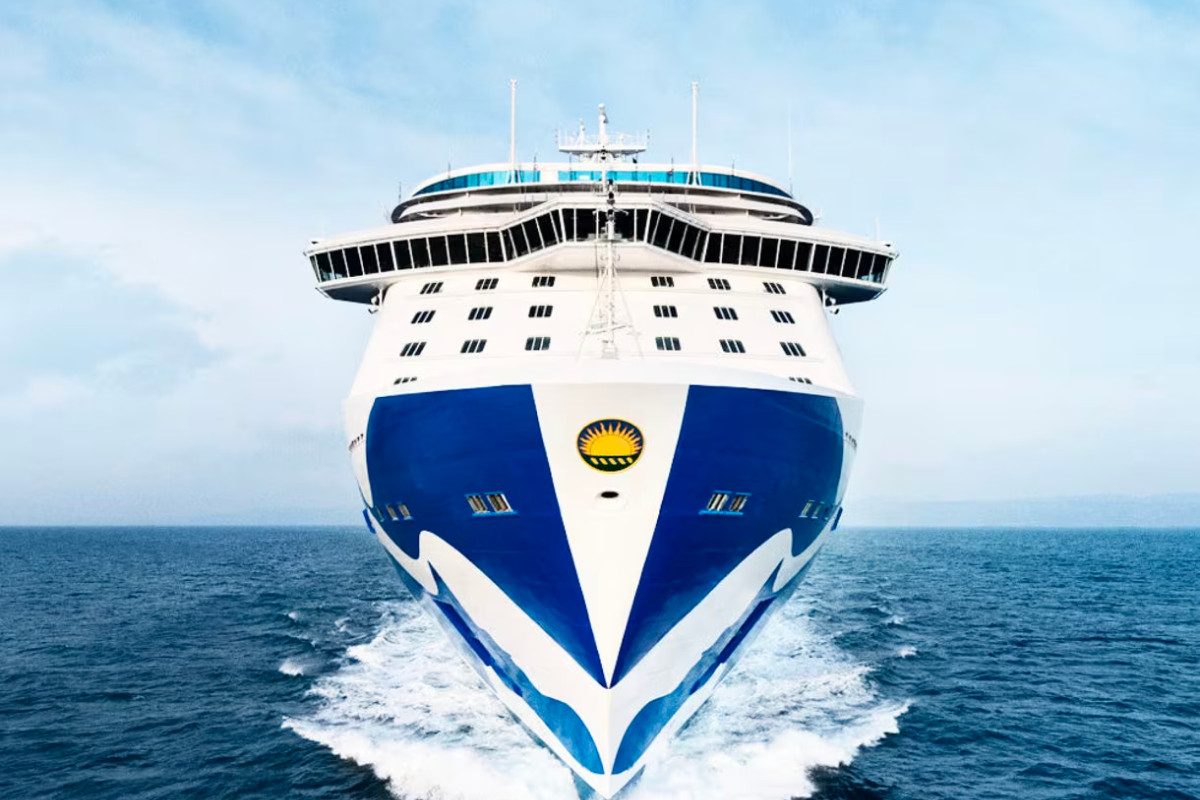 A front-facing view of a large Princess Cruises ship cutting through the ocean. The ship's bow, painted in blue and white with the iconic Princess Cruises logo, dominates the image as it moves steadily through the water under a bright blue sky. The vast sea stretches out behind the vessel.