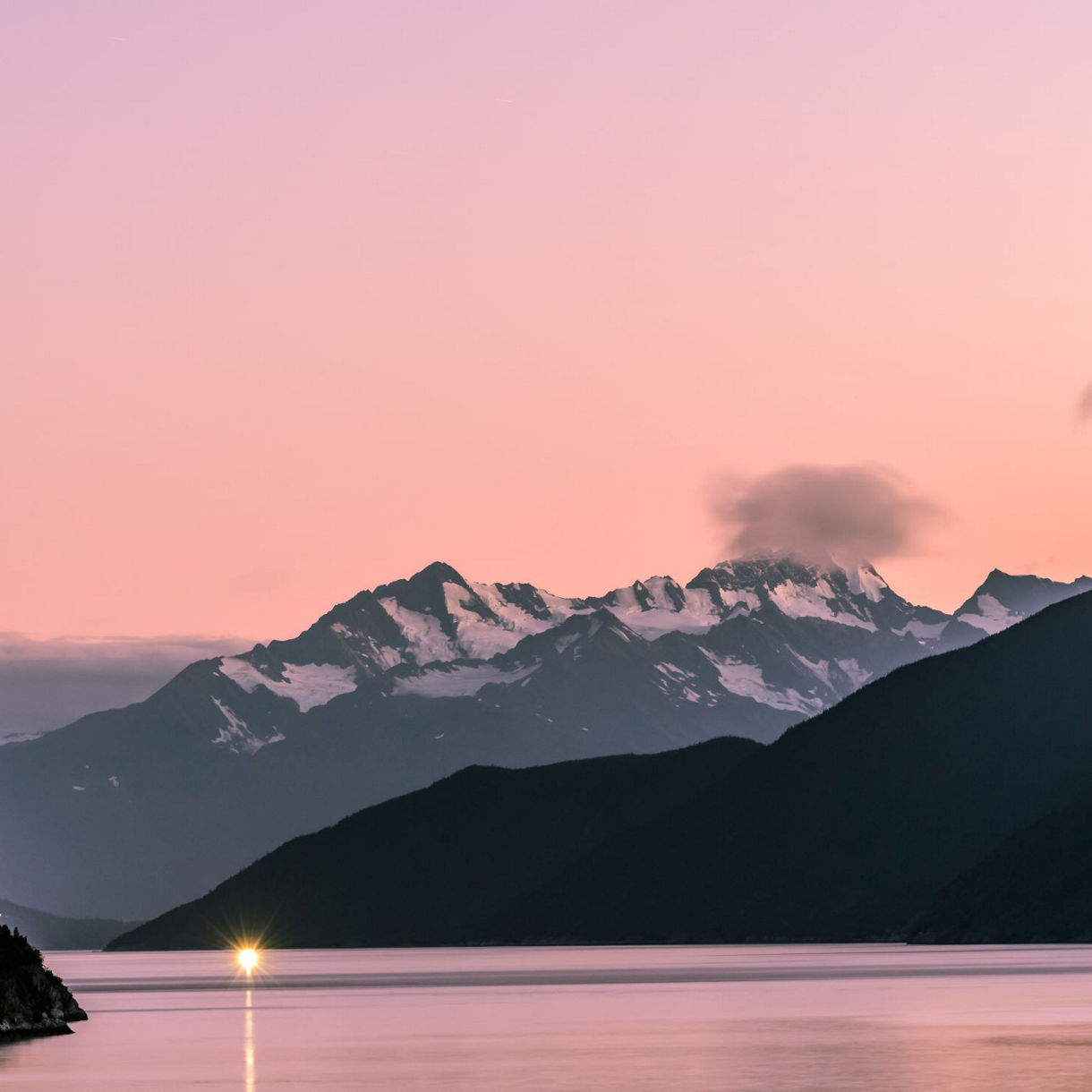A picturesque sunset in Skagway, Alaska, with vibrant shades of orange and pink illuminating the sky, silhouetting the surrounding mountains, and reflecting on the peaceful waters below.