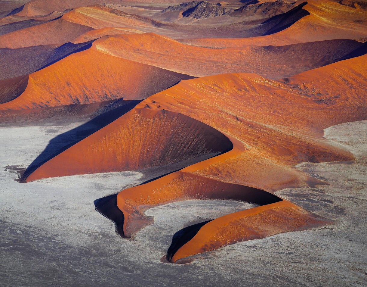 Aerial view of Namibia’s Sossusvlei dunes showing sweeping orange sand ridges, deep shadows and pale salt flats creating striking patterns across the desert landscape.