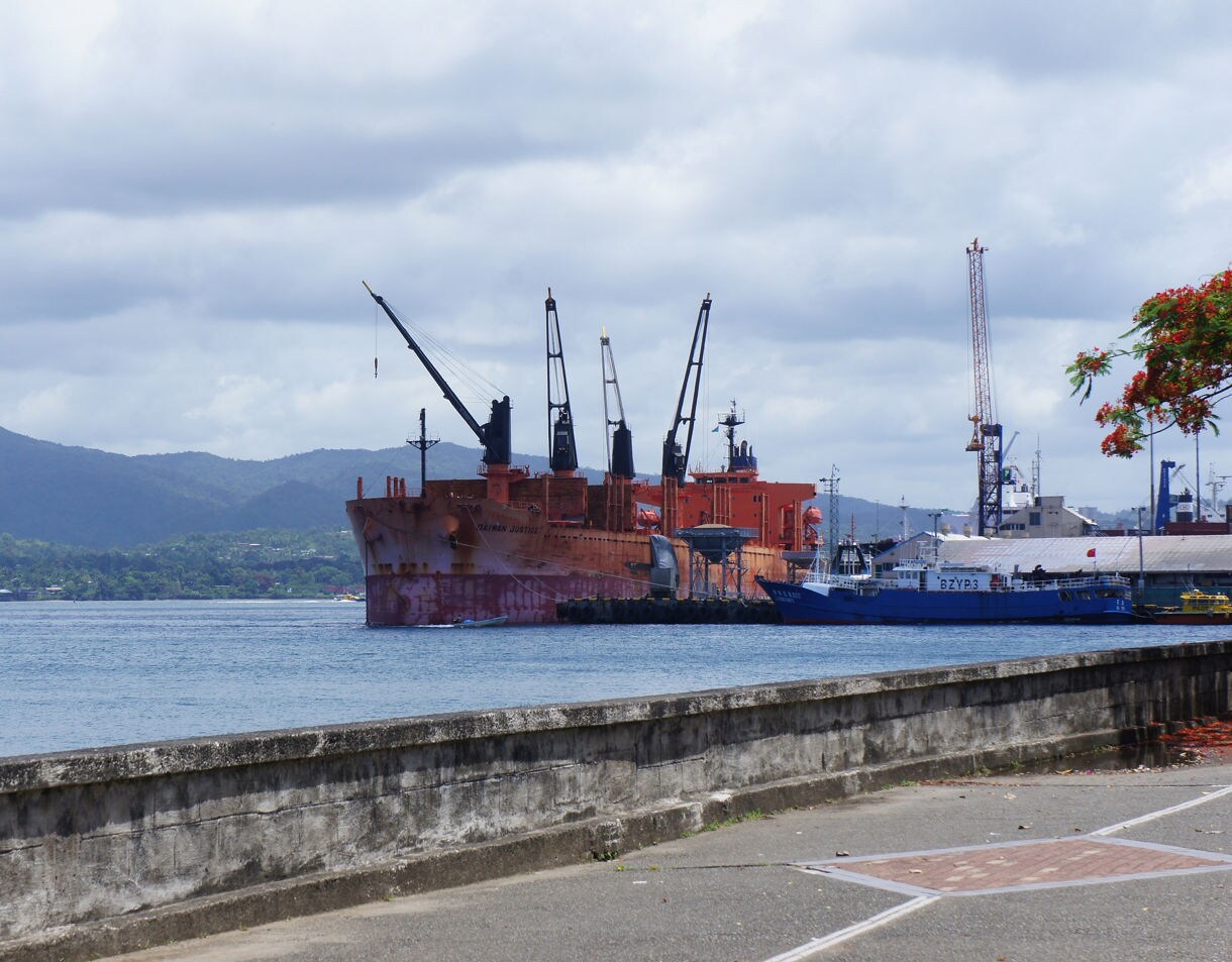 A rust-colored cargo ship and a smaller blue vessel docked in Suva harbor with forested hills in the background under cloudy skies.