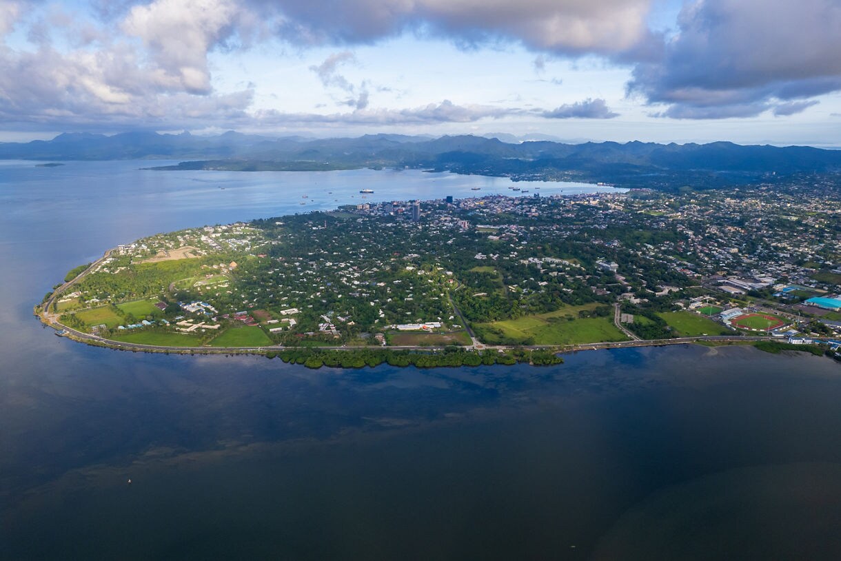 Aerial view of Suva’s coastline in Fiji, showing green neighborhoods, a curving shoreline, calm ocean water and blue mountains in the distance under scattered clouds.