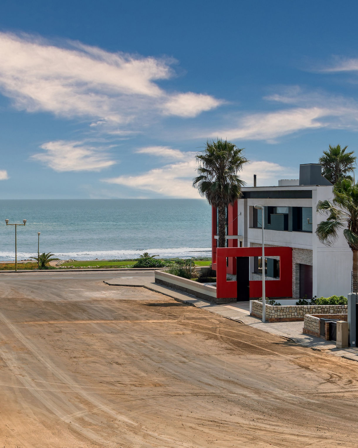 View of a sandy residential street in Swakopmund leading to the ocean, with tall palm trees, modern houses and gentle waves visible beneath a blue sky with soft clouds.