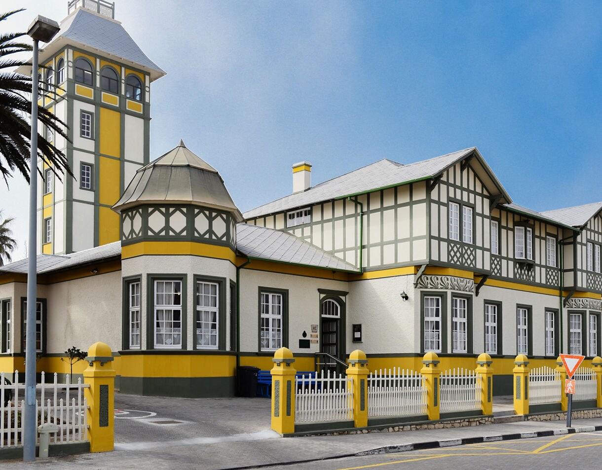 Historic German colonial-style building in Swakopmund with green and yellow trim, white picket fencing, a tall tower and multiple windows, all set beneath a bright blue sky.