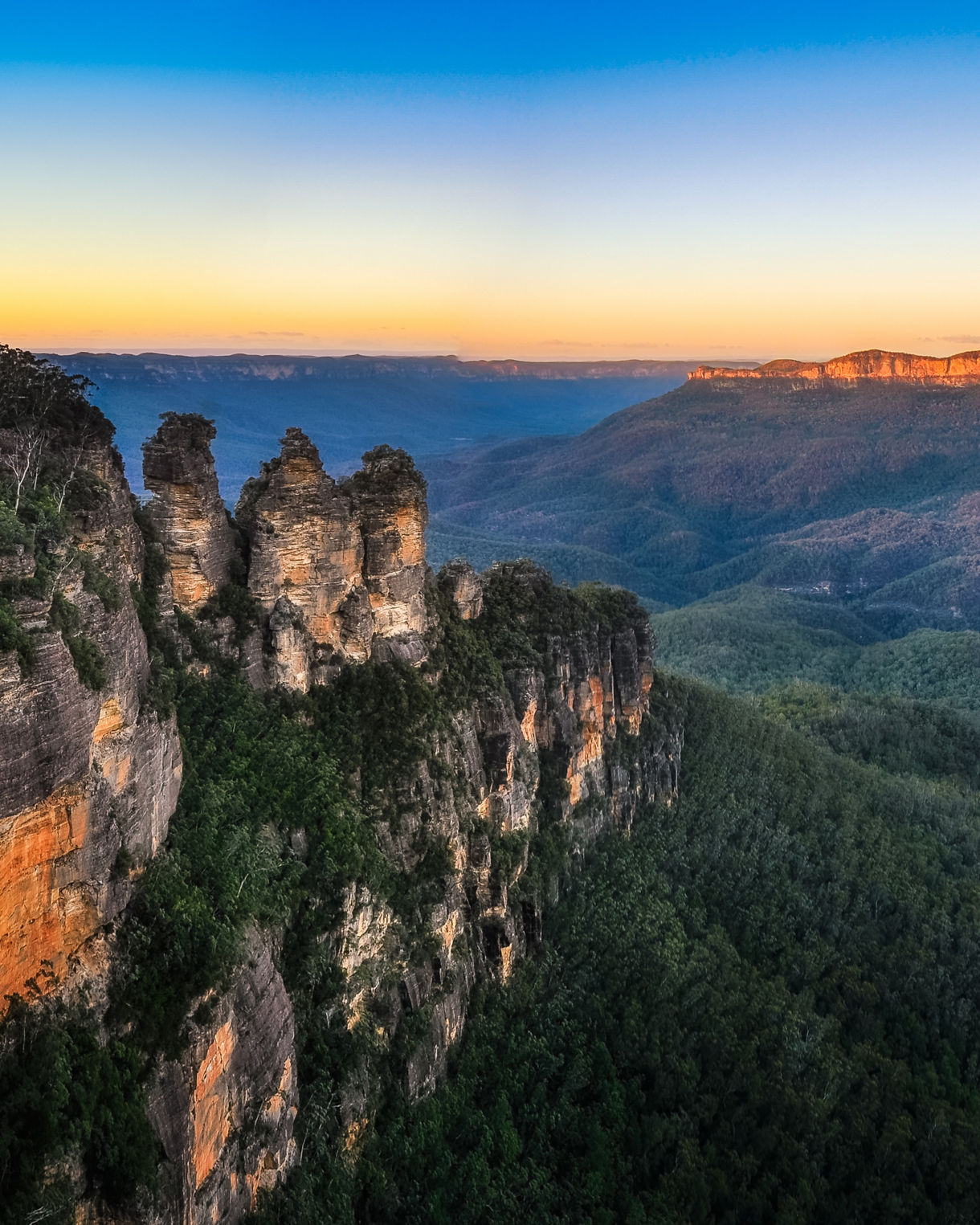 Panoramic view of the Blue Mountains featuring the Three Sisters rock formation on a rugged cliff, overlooking rolling forested valleys under a clear sky lit by soft early morning light.