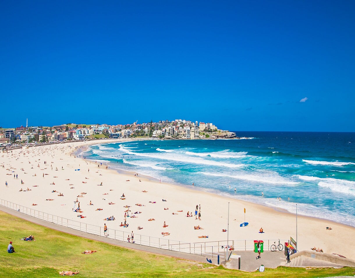 Wide view of Bondi Beach with bright turquoise waves, a long stretch of white sand filled with people sunbathing, and a cluster of coastal buildings rising above the curve of the bay.
