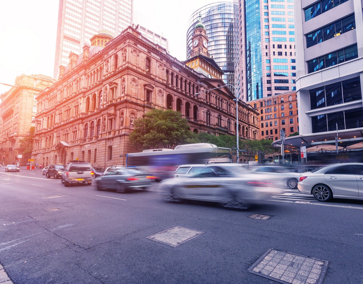 Street-level view of downtown Sydney with blurred cars moving through a busy intersection, framed by a grand historic sandstone building with a clock tower and surrounding modern skyscrapers.