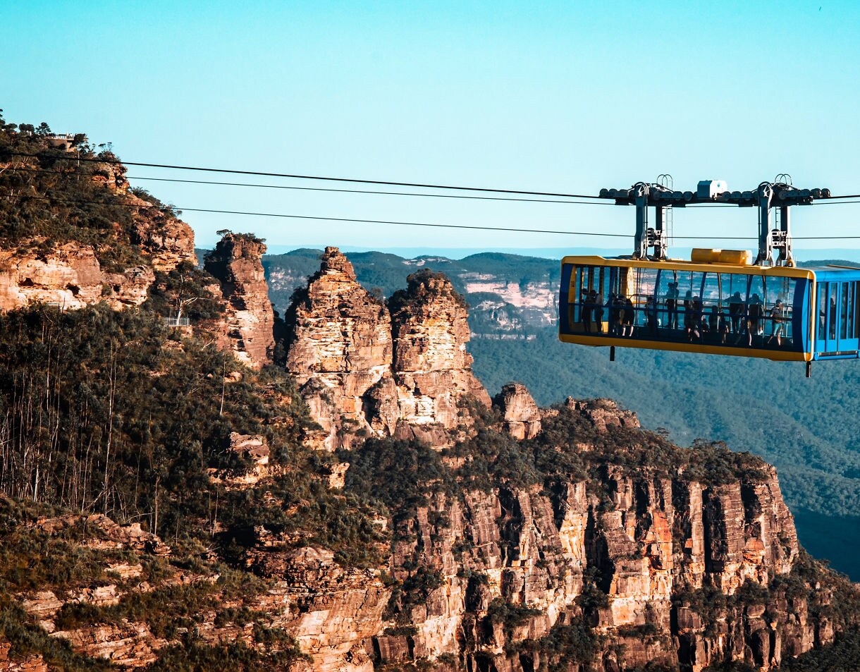 A bright blue and yellow cable car suspended over the Blue Mountains, with passengers inside and rugged sandstone cliffs and forested valleys stretching into the distance.