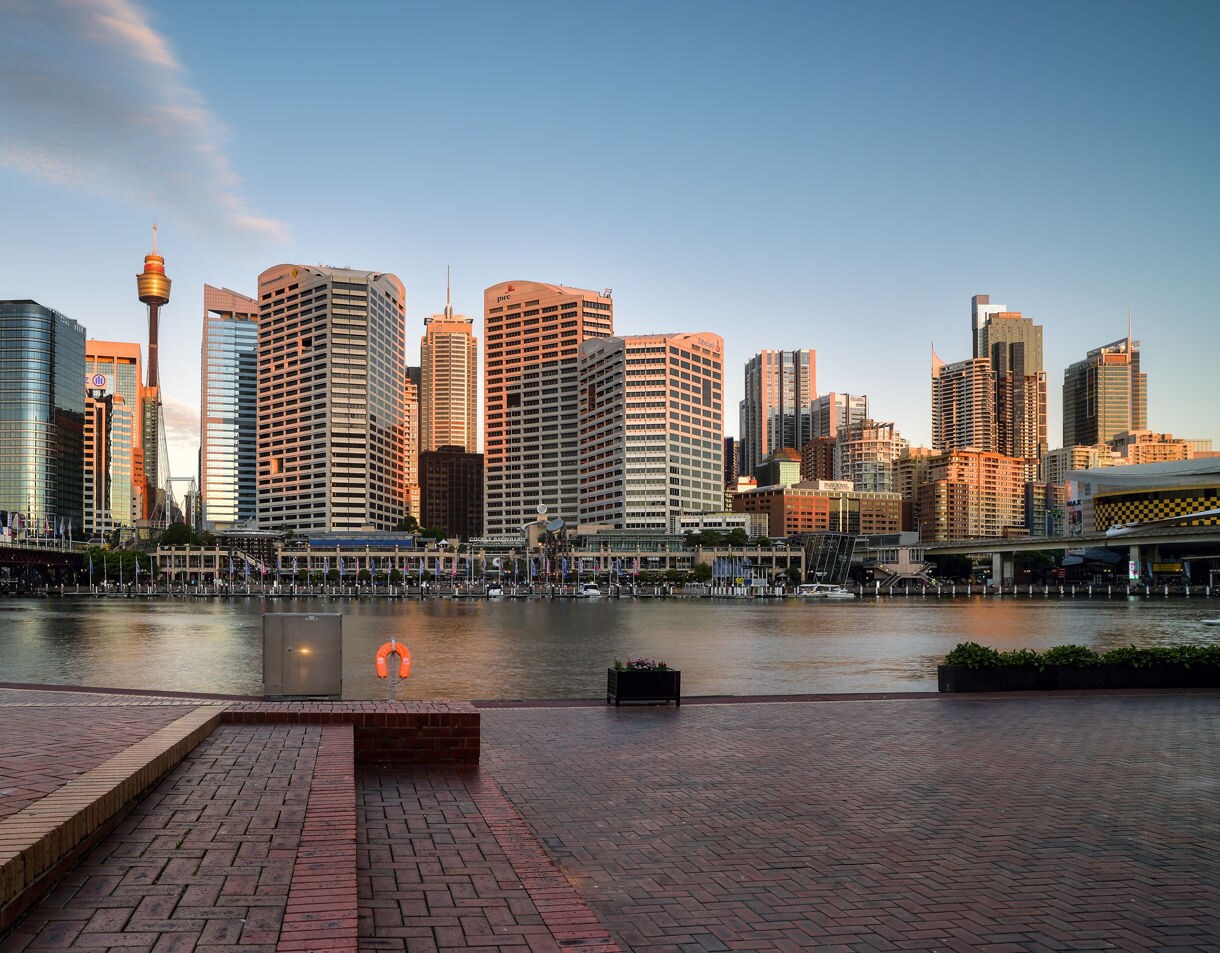 Waterfront view of Darling Harbour with tall modern buildings glowing in soft sunset light, a brick promenade in the foreground and calm water reflecting the cityscape.