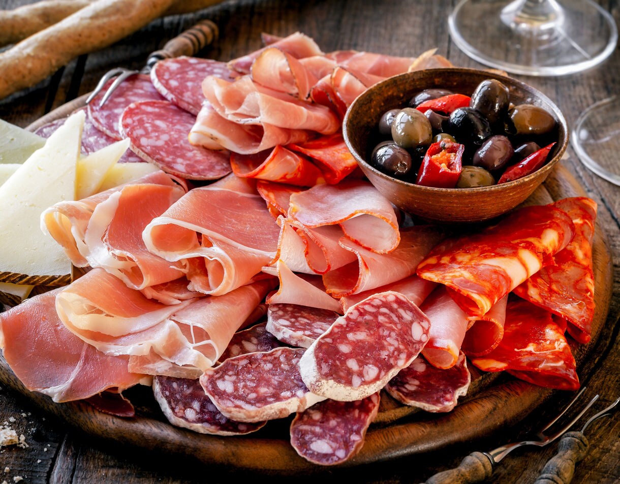 Close-up of a charcuterie board featuring sliced prosciutto, salami, chorizo, wedges of firm cheese and a small bowl of mixed olives with peppers, all arranged on a rustic wooden platter.
