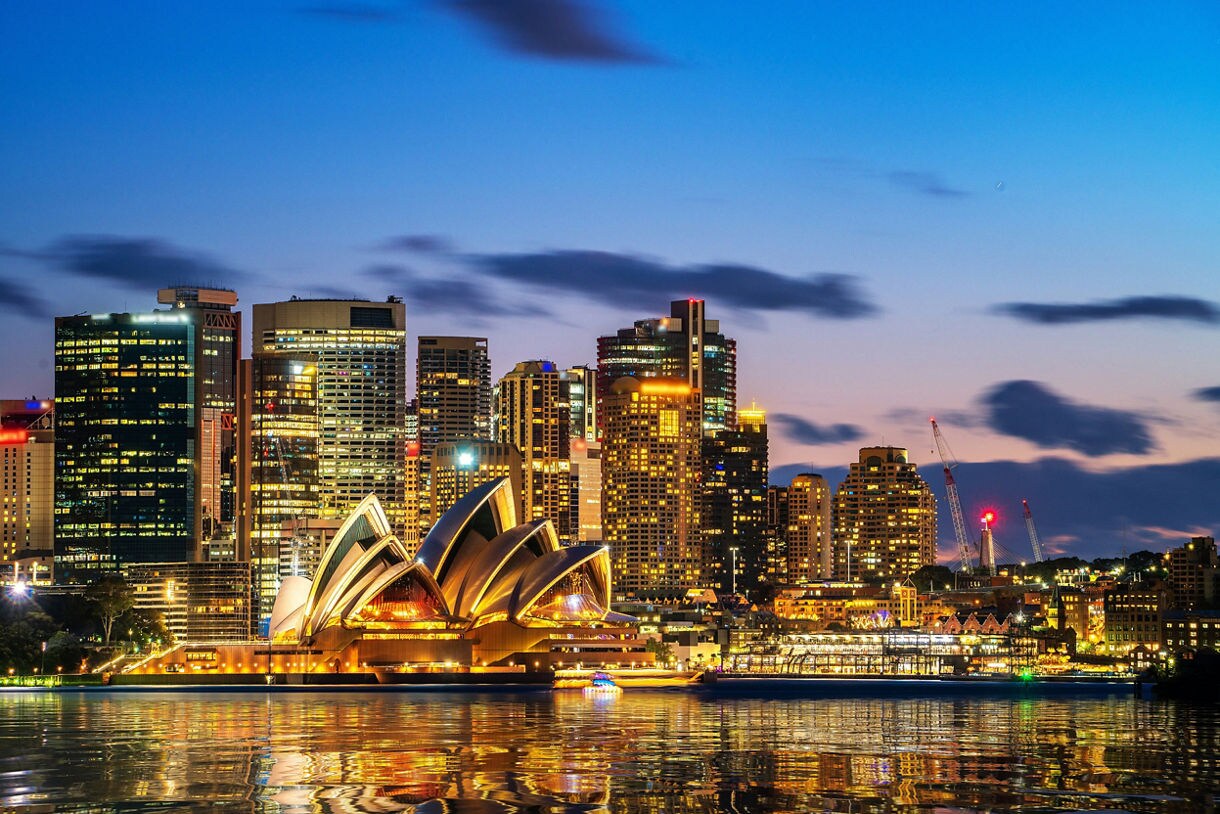 Night view of the Sydney Opera House illuminated in warm gold tones with the city skyline rising behind it, reflected across the calm harbor waters under a deep blue sky.