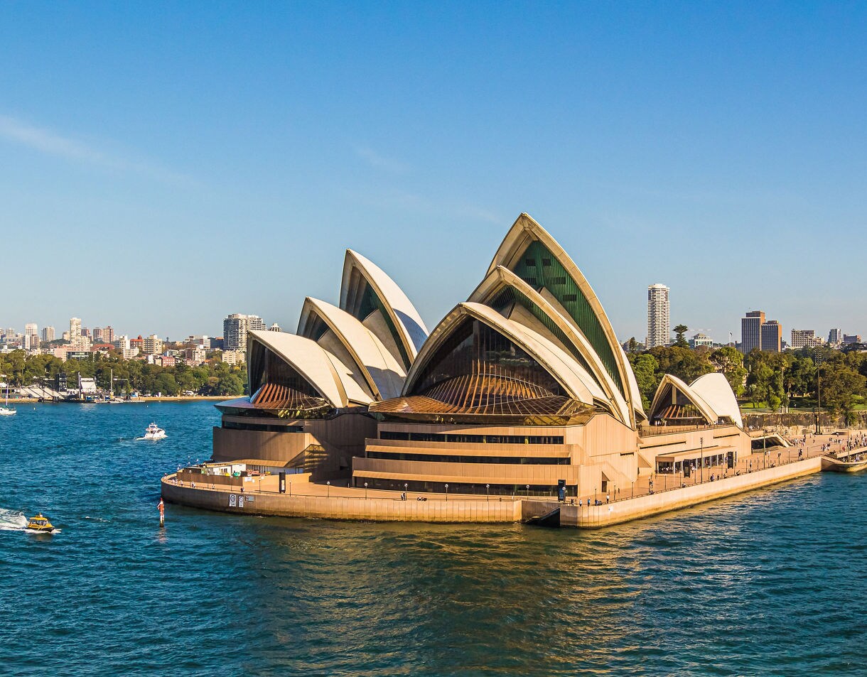 Bright daytime view of the Sydney Opera House with its white sail-like roofs, set beside deep blue harbor water and backed by city skyscrapers and green parkland.