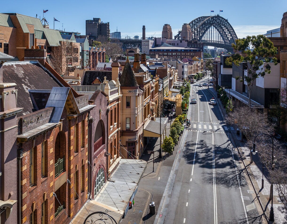 Elevated view of The Rocks district in Sydney featuring historic brick buildings, a quiet street lined with shops and trees, and the Sydney Harbour Bridge rising in the background under a clear blue sky.