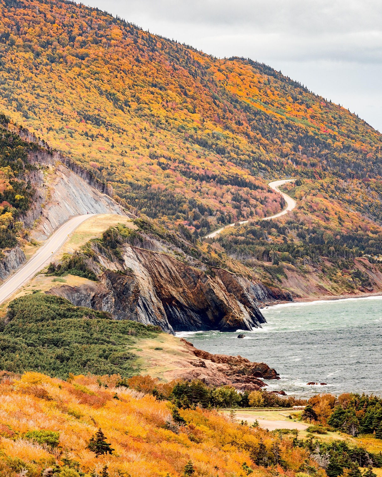 Vertical view of the Cabot Trail running along steep cliffs above the ocean, surrounded by dense fall foliage in orange, yellow and red tones under an overcast sky.