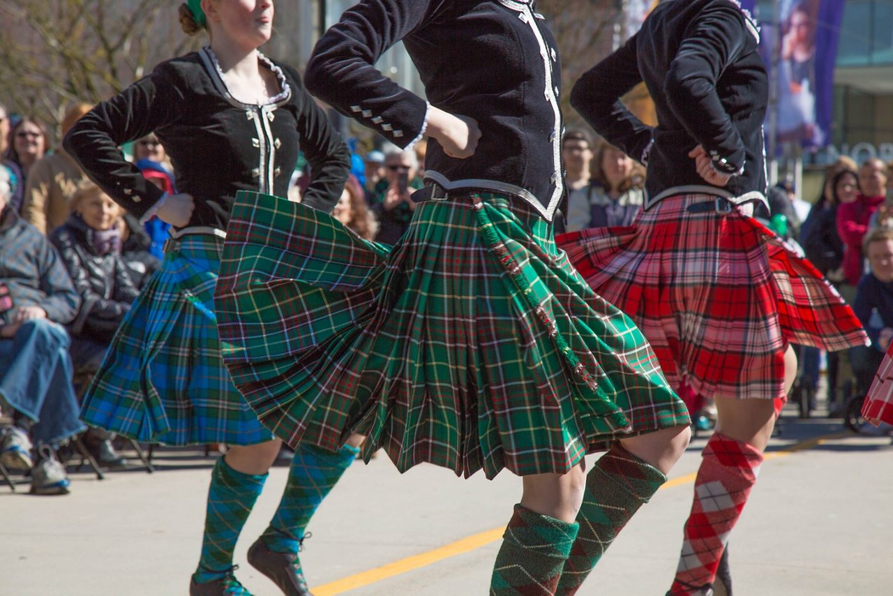 Three dancers in colorful tartan kilts and matching knee-high socks performing a traditional Celtic dance outdoors, with their skirts flaring as they move and a seated crowd watching in the background.