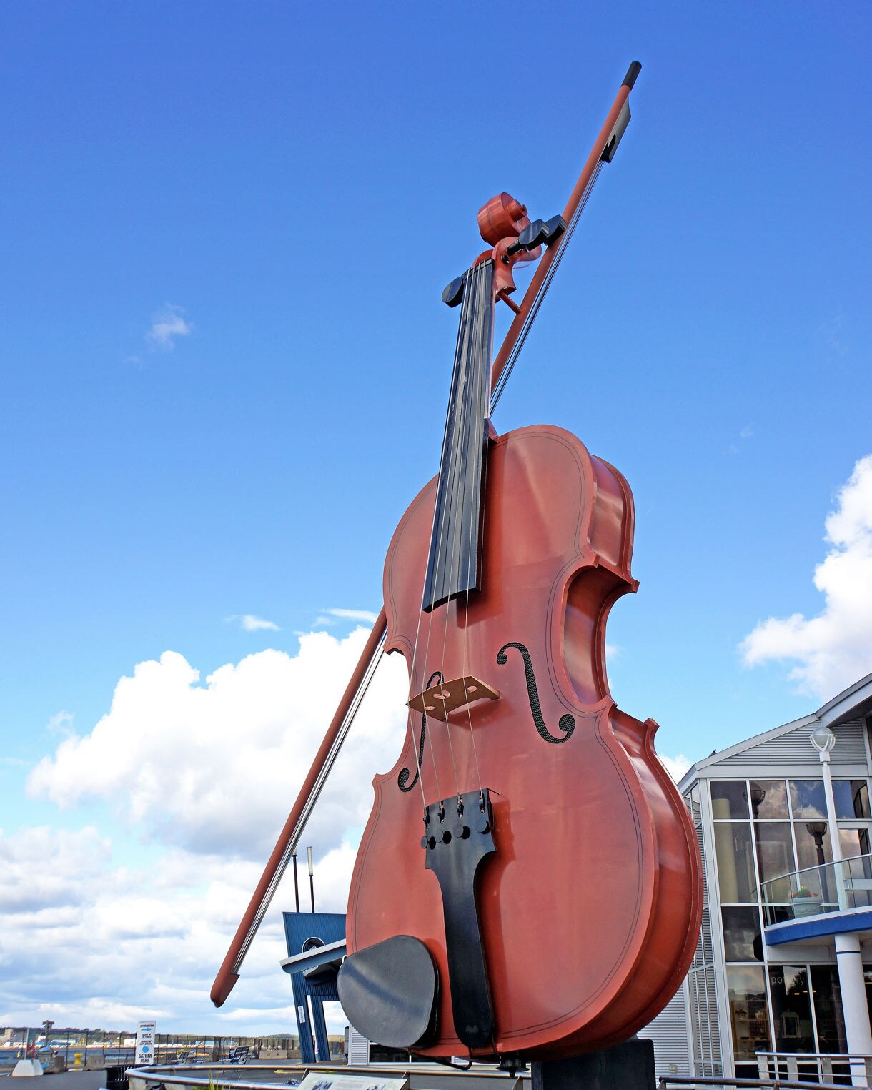 A massive outdoor fiddle sculpture with an attached bow rising several stories high, set along a waterfront walkway with benches, railings and a modern building nearby under a bright blue sky with scattered clouds.