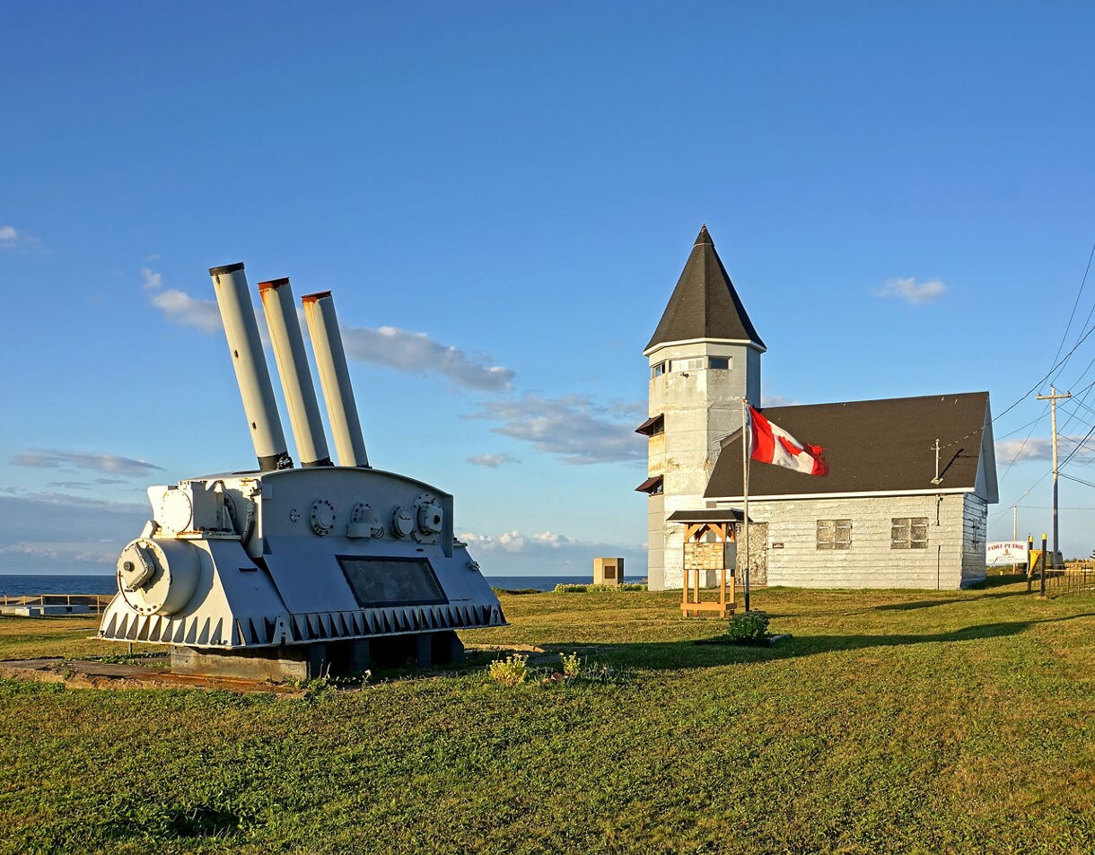 An old coastal defense site featuring a grey naval gun mount with three barrels on the left and a worn wooden tower building flying a Canadian flag on the right, all set on a grassy hill beside the ocean under a clear blue sky.