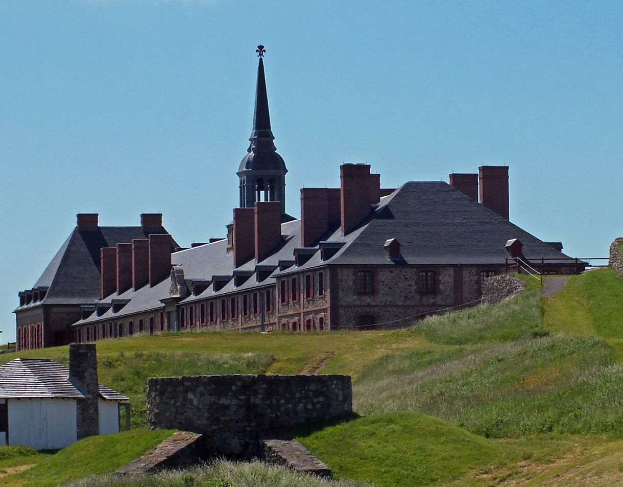 Historic stone buildings and tall chimneys of Fortress Louisbourg set behind grassy embankments and fortified walls, with a central steeple rising into a clear blue sky.
