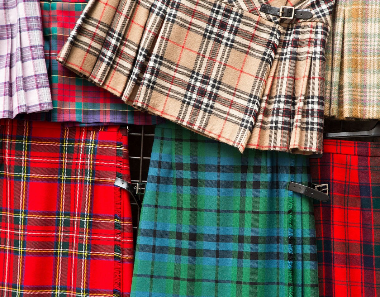 Close-up view of assorted tartan fabrics in red, green, blue, purple and beige patterns, arranged in rows with pleats and buckled straps visible on several kilts.