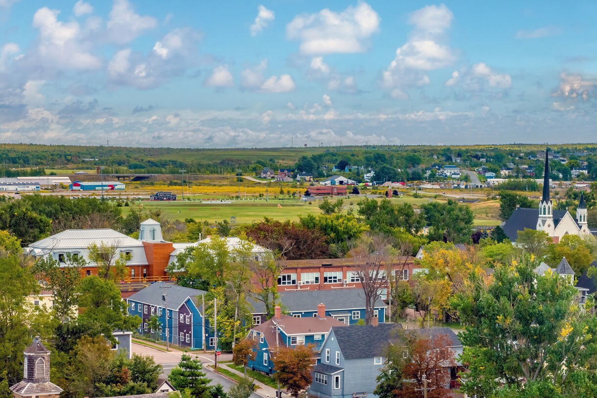A panoramic view of a small Canadian town with blue and gray houses, leafy trees and a large church with tall steeples on the right, set against sprawling fields, low buildings and distant rolling hills beneath a bright blue sky with scattered clouds.
