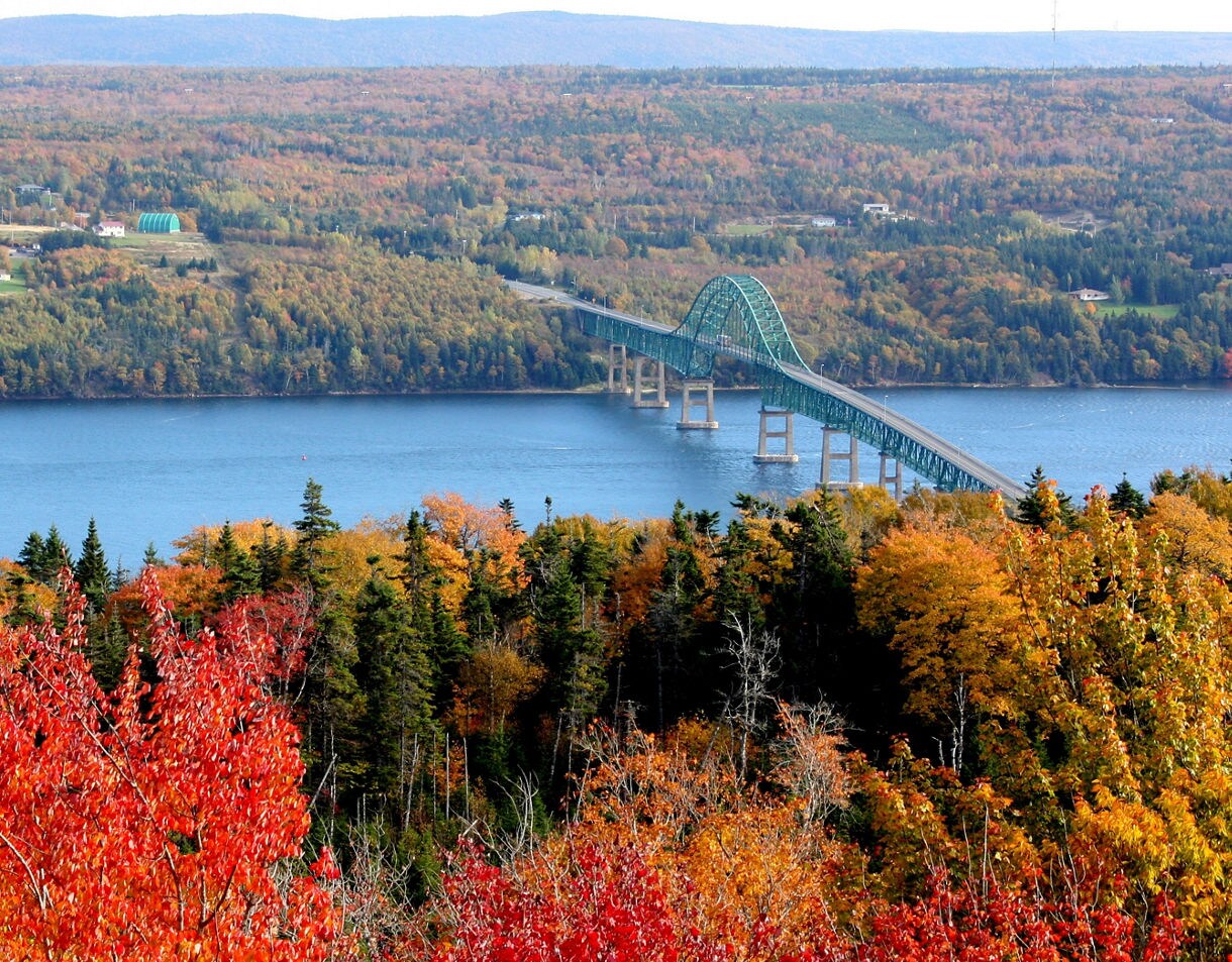 Scenic view of Seal Island Bridge spanning a wide blue channel, surrounded by dense forests in peak fall colors of red, orange and yellow, with rolling hills fading into the distance.