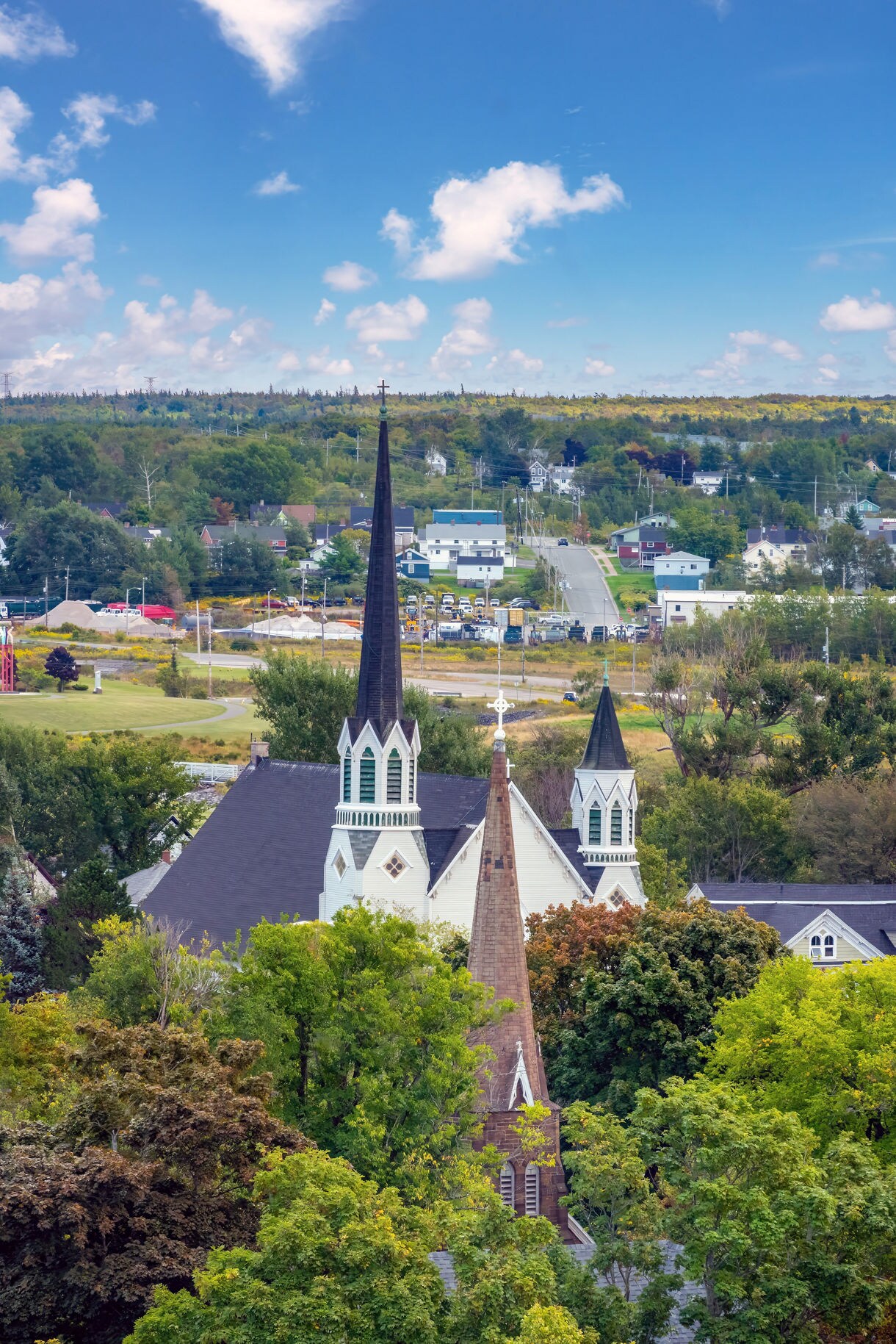 Elevated view of a small Canadian town featuring tall church steeples surrounded by dense green trees, with houses, roads and rolling wooded hills stretching into the distance under a bright blue sky with scattered clouds.