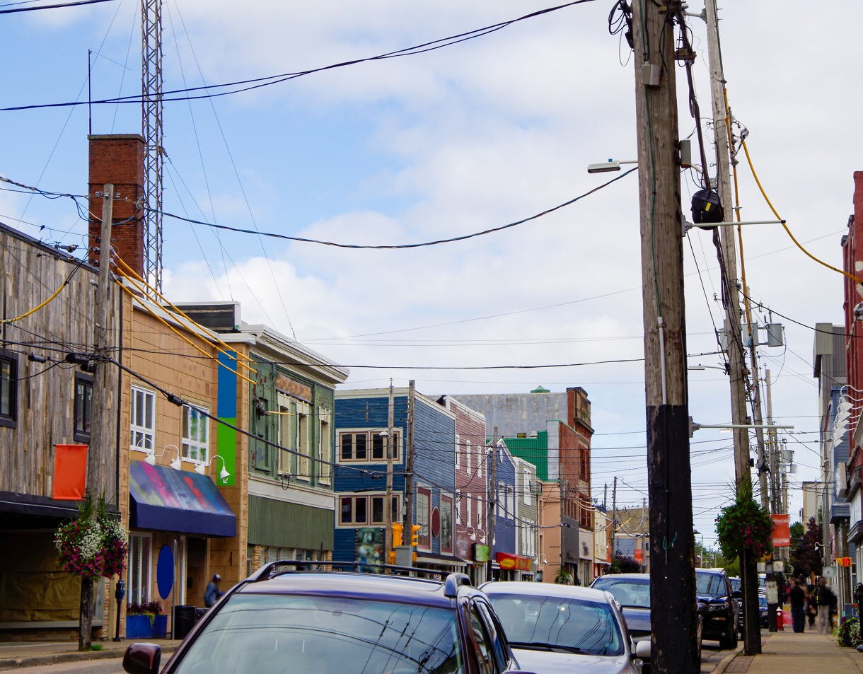 Urban street scene featuring shops with painted facades, parked cars, hanging flower baskets and numerous overhead power lines under a partly cloudy sky.
