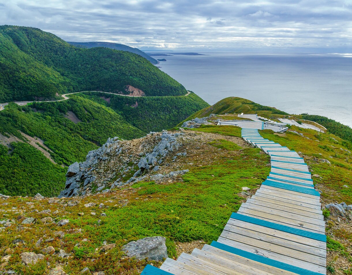A curving wooden boardwalk with turquoise trim leads across a rocky, grassy ridge overlooking forested mountains and a calm ocean under a cloudy sky.