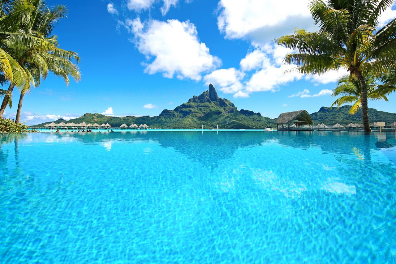 A stunning view of a resort in Bora Bora, French Polynesia, featuring crystal-clear turquoise waters, overwater bungalows, palm trees, and the iconic jagged peak of Mount Otemanu in the background.