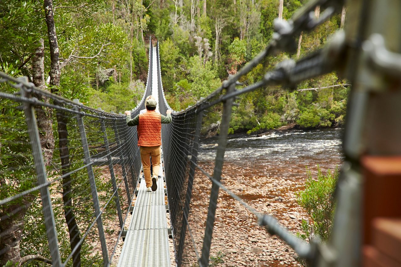 Hiker crosses suspension bridge over rocky river. 