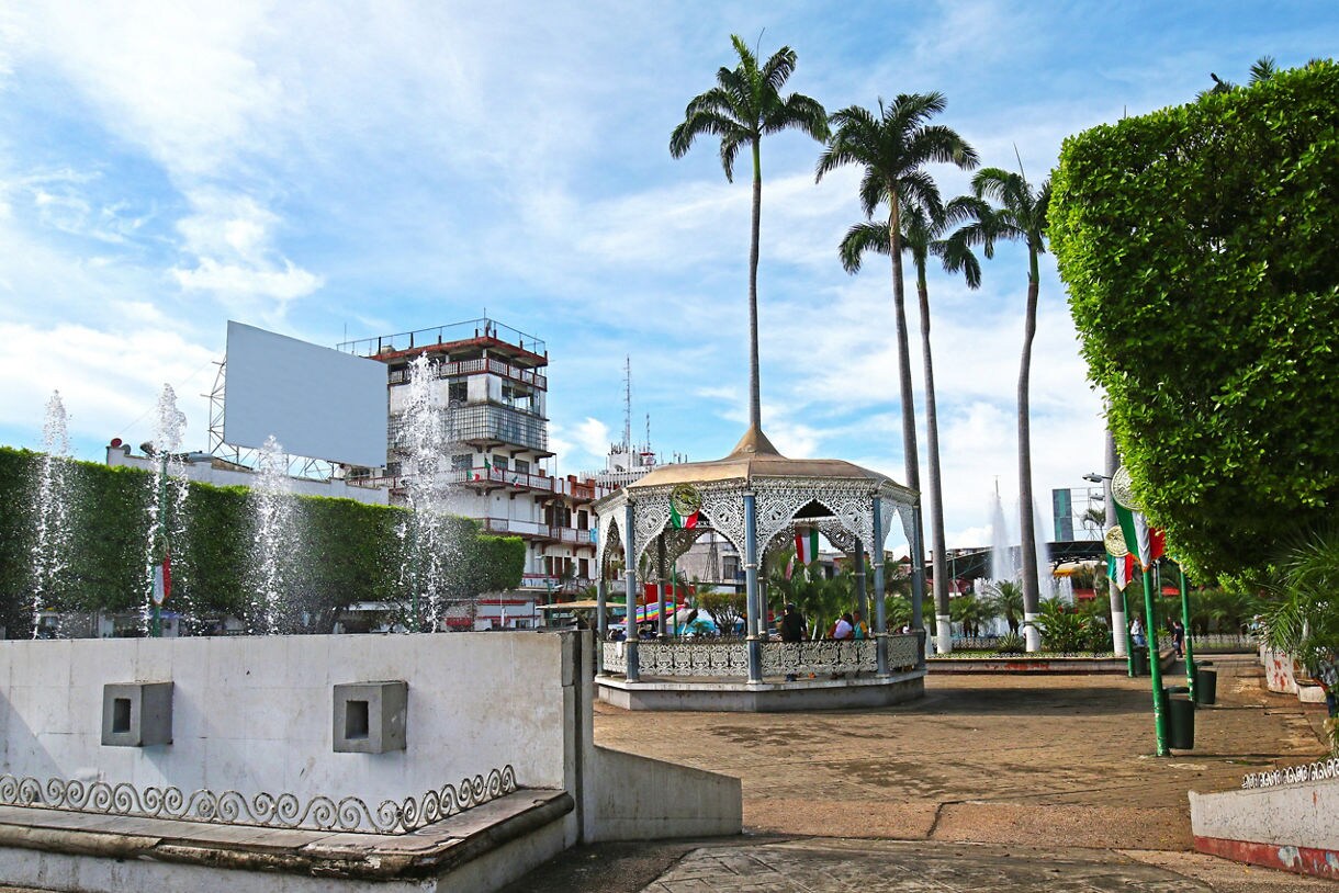 Public square in Tapachula, Chiapas, with a decorative iron gazebo, tall palm trees, fountains and buildings in the background decorated with Mexican flags.