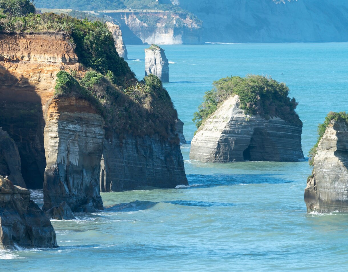 Tall layered sea cliffs and isolated rock stacks covered with greenery standing in turquoise coastal waters near Taranaki, New Zealand.