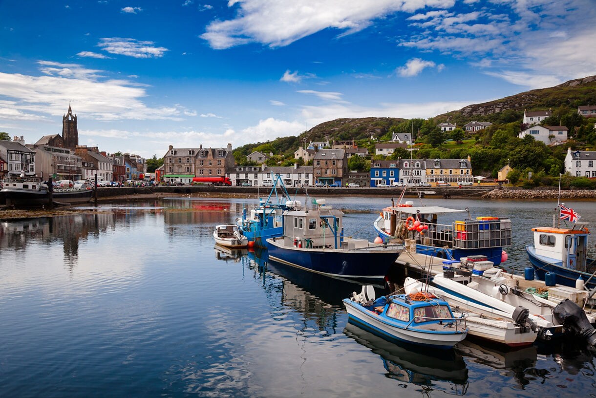 Fishing boats tied to a quiet harbor in Tarbert with stone buildings, hillside homes and blue sky reflected in the calm water