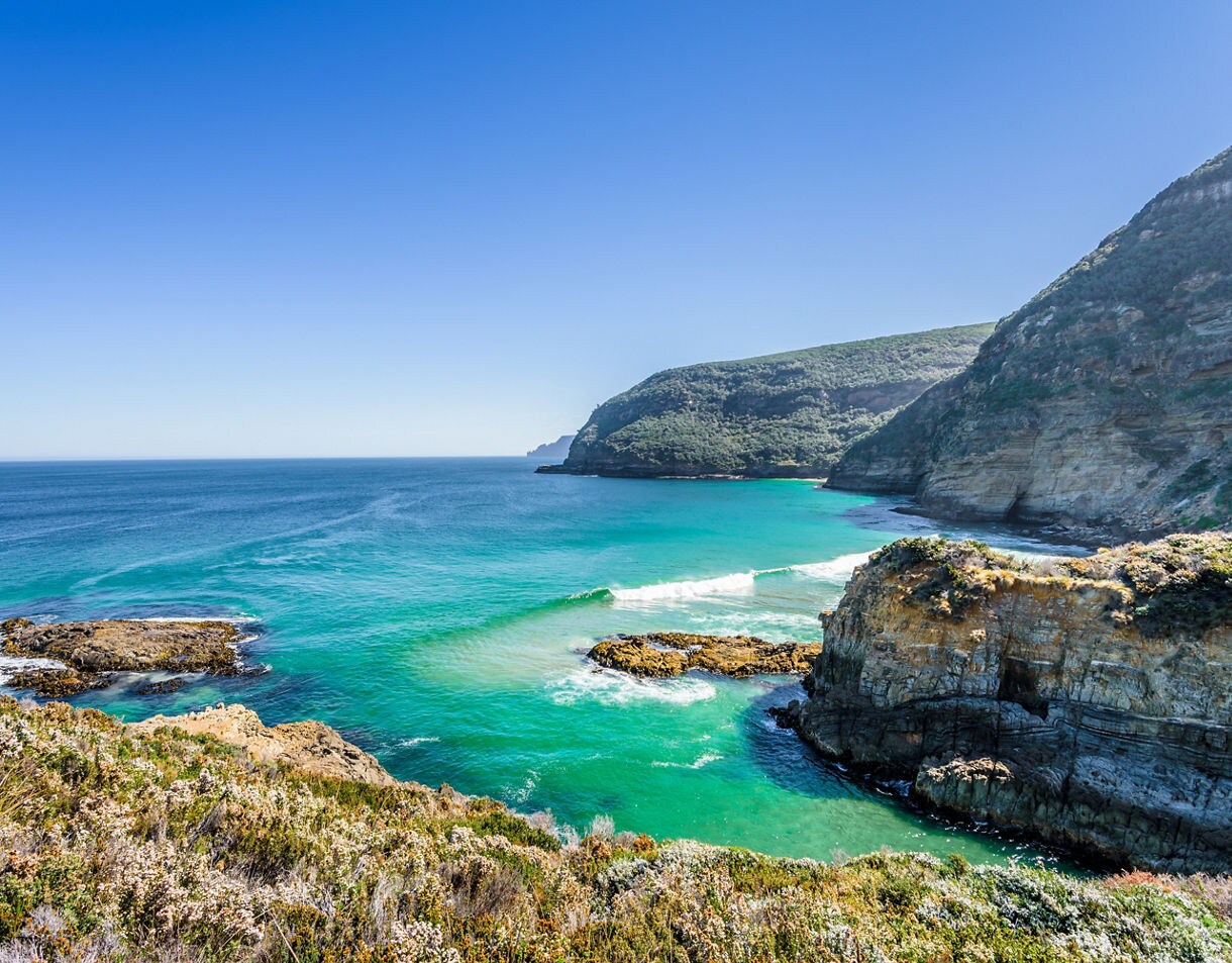 View of vibrant turquoise ocean waters along rocky coastal cliffs on the Tasman Peninsula, with waves breaking near shore and hills covered in greenery under a clear blue sky.