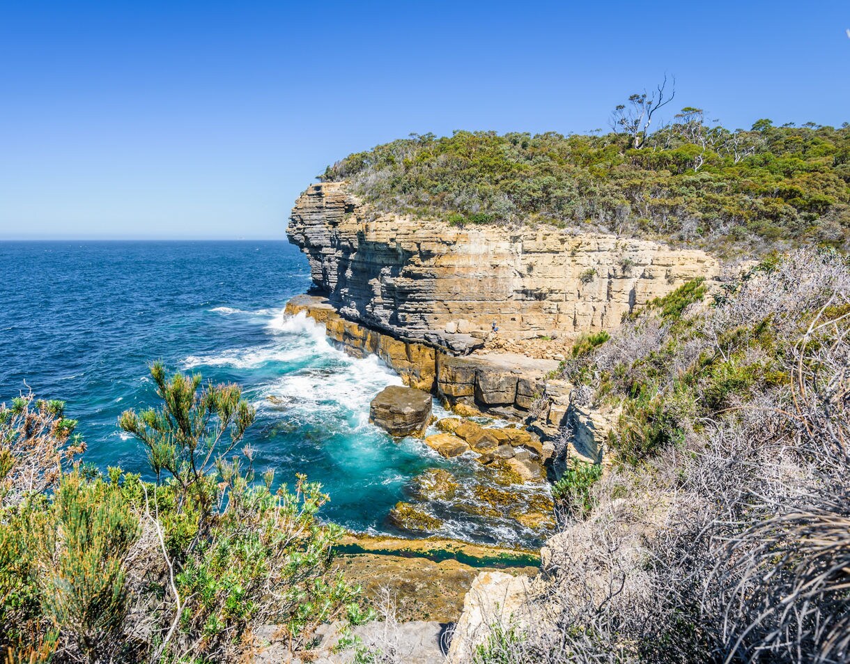 Rocky coastal cliffs with turquoise waves swirling below, framed by scrubby coastal vegetation under a bright blue sky.