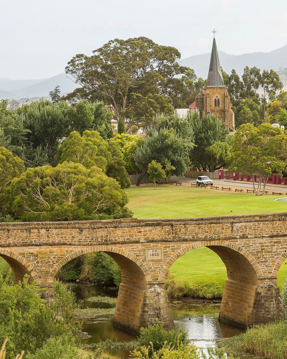 Historic stone bridge crossing a small river with a church steeple and leafy village behind it.
