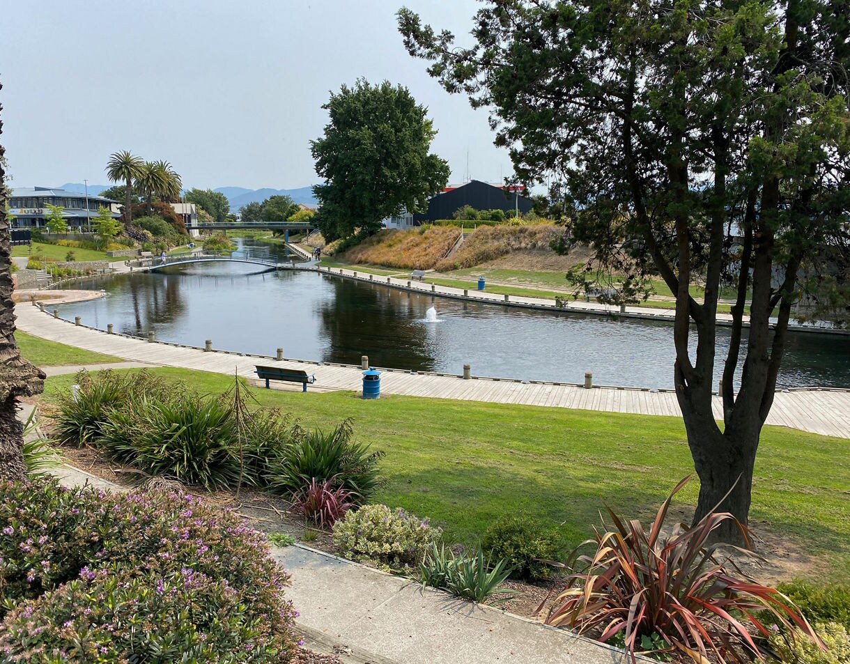 A quiet river bordered by grassy banks, a wooden boardwalk, trees and a bench, with a small bridge and low hills in the distance.