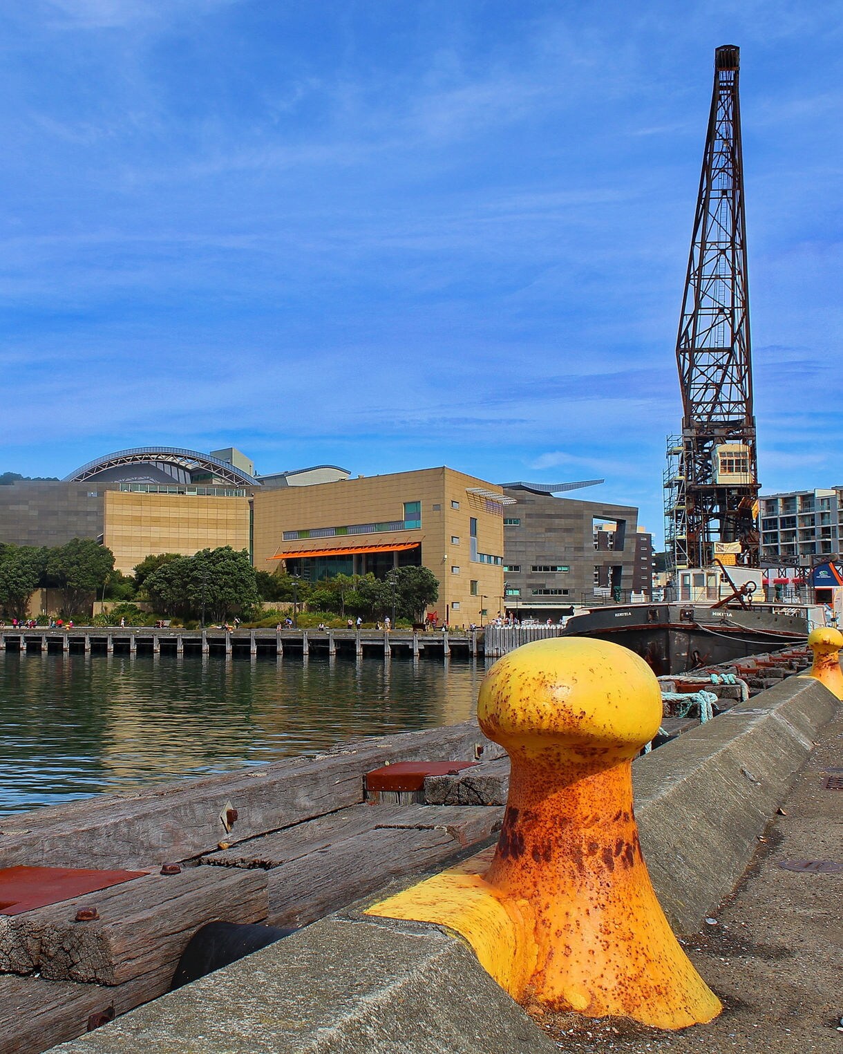 View of Te Papa Tongarewa museum along Wellington’s waterfront, with a dockside crane, yellow mooring posts and people walking by the harbor.