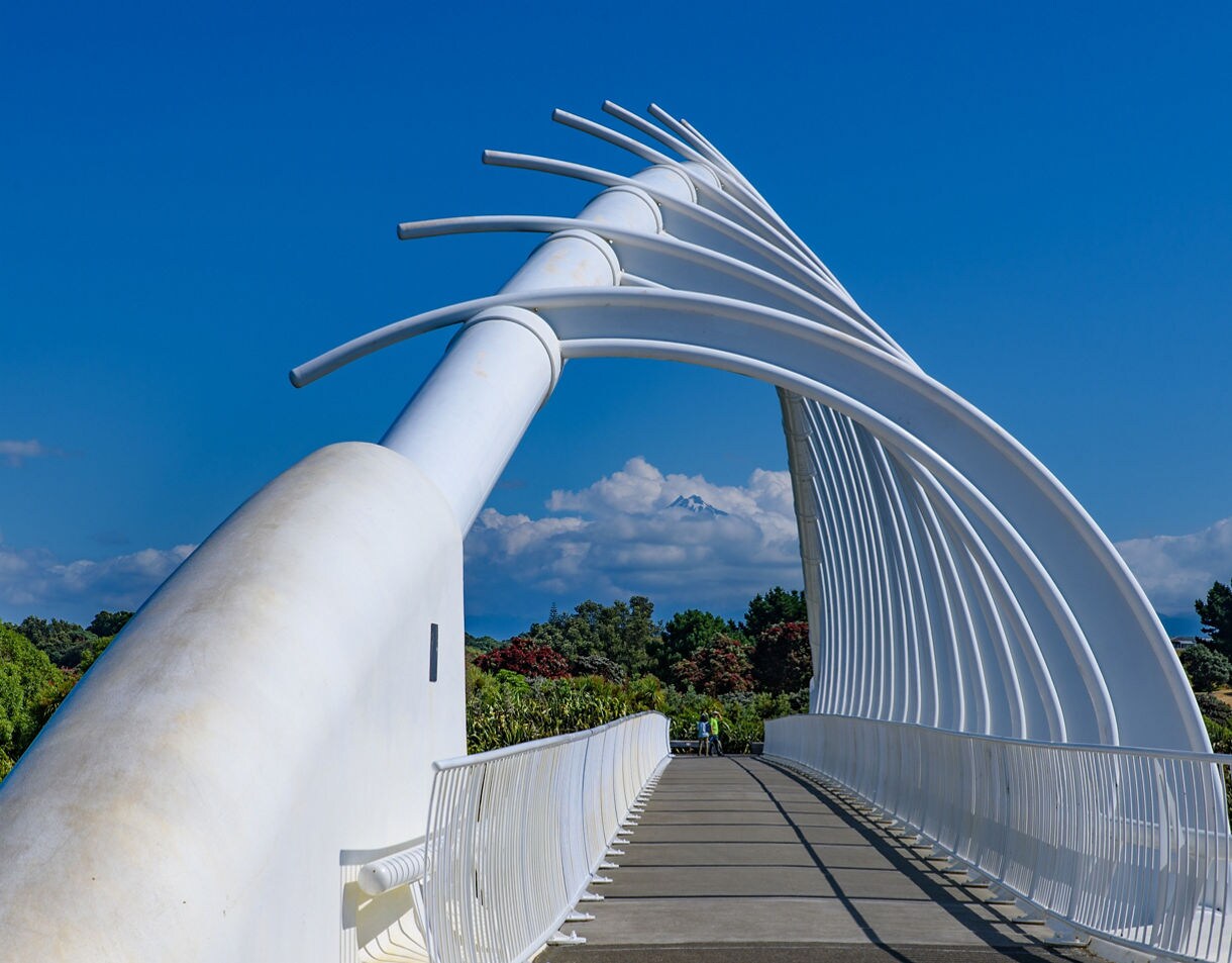 A modern white sculptural bridge with curved rib-like arches, leading toward lush greenery and a distant snow-capped Mt Taranaki under a bright blue sky.
