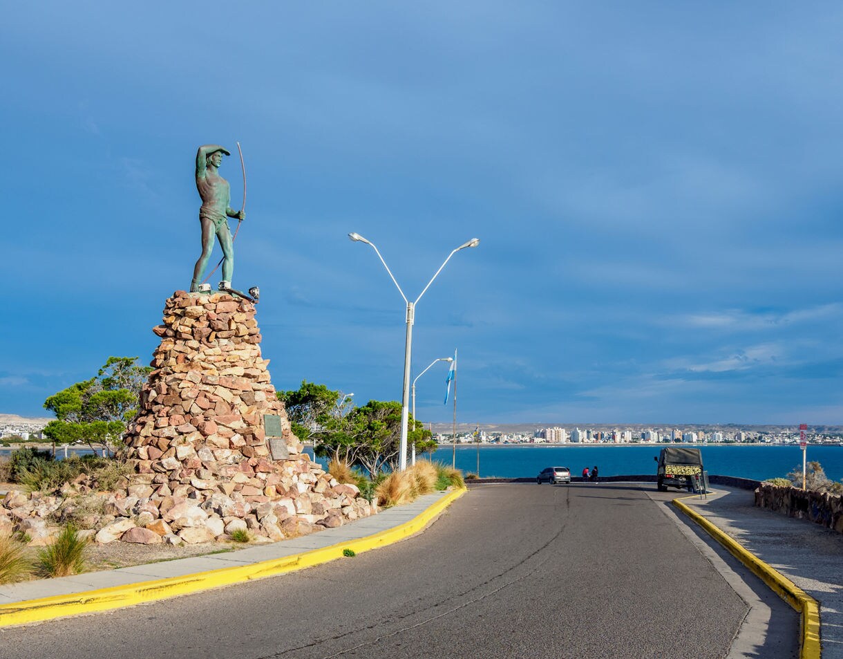Coastal road leading to Puerto Madryn with a tall stone base topped by a bronze Tehuelche figure holding a bow, set against a vivid blue sky and calm ocean.