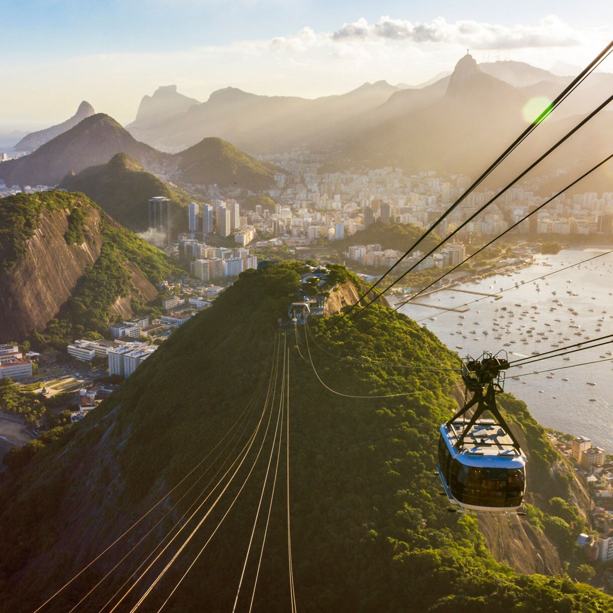 Aerial view of a mountainous landscape in South America, showcasing lush greenery and a cityscape in the background, with a cable car traveling down a steep hill towards the ocean. 