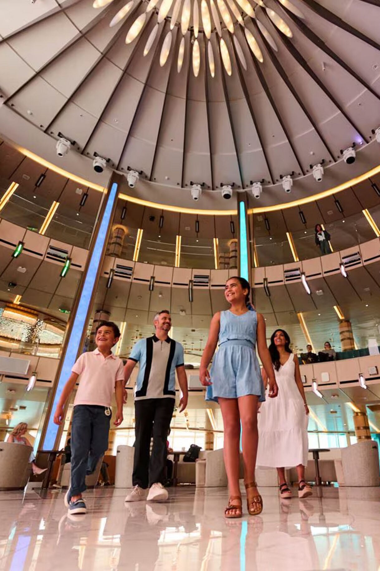 Guests walking through a modern cruise ship atrium featuring a distinctive circular skylight with radiating panels and polished marble floors.