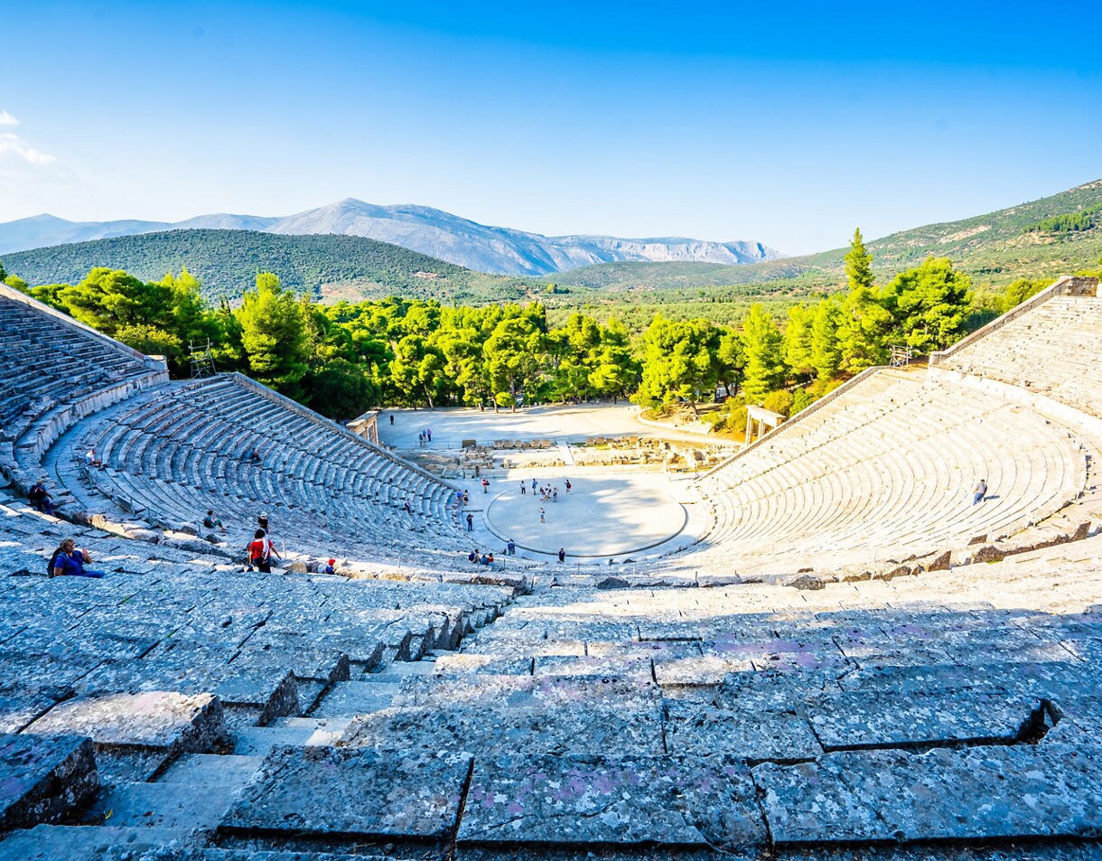 A wide view of the ancient Theatre of Epidaurus in Greece, showing its curved stone seating set amid green hills and mountains under a clear blue sky.