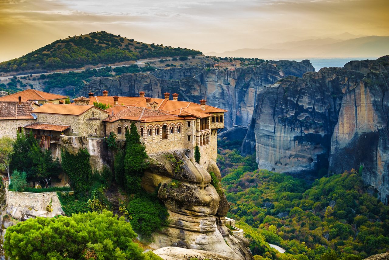 A historic monastery perched on the edge of a rocky cliff at Meteora, Greece, with dramatic mountainous landscapes and lush green valleys in the background.